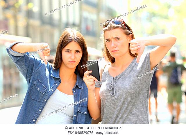 Two angry friends holding a smart phone with thumbs down in the street