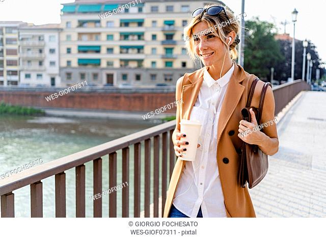 Smiling woman with earphones and coffee to go walking through city