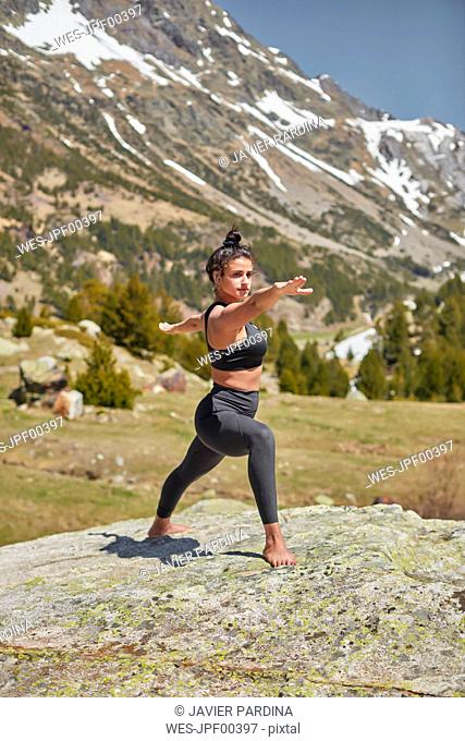 Young woman doing yoga standing on rock in nature, warrior pose