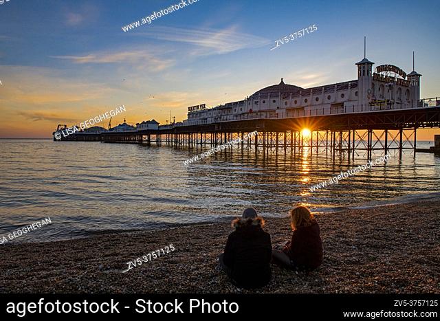 Two girls watch the sun setting at the Palace Pier, Brighton, East Susees, England, Uk