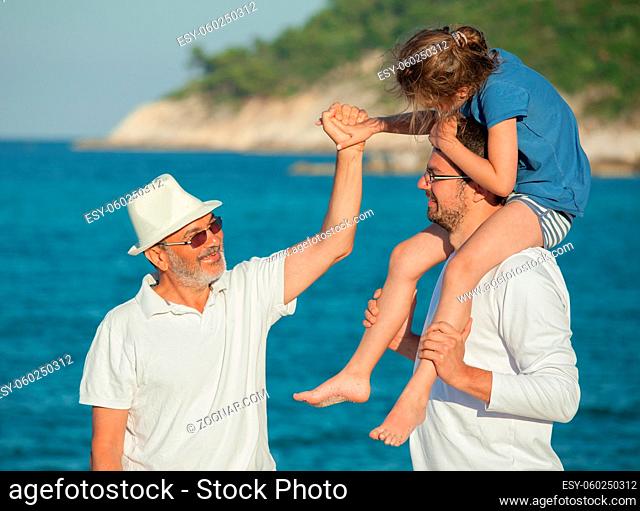 Grandfather holding girl s hand while she is riding her father s shoulders on a sea beach