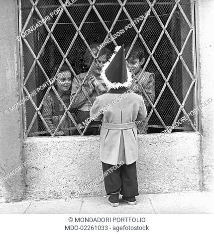A child wearing a carnival hat talking with some children behind a grating. Bergamo, 1950s