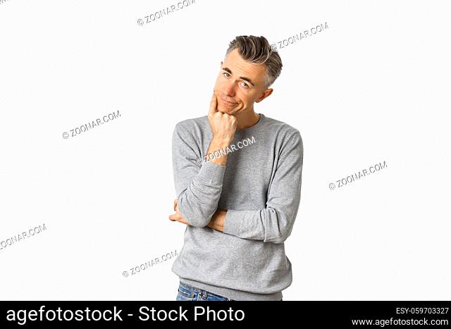 Portrait of bored and unamused middle-aged man in grey sweater, leaning on hand and looking at camera reluctant, standing sad over white background