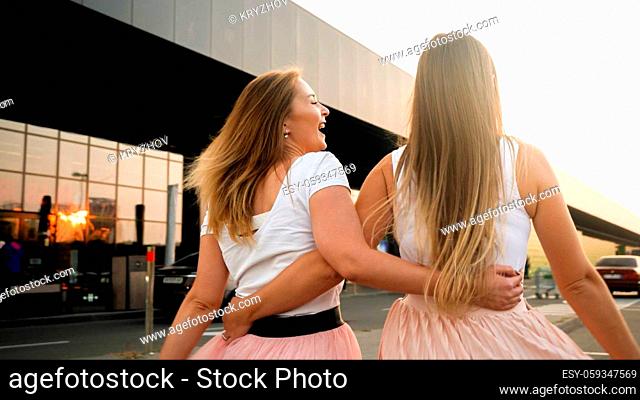 Rear view photo of two young women laughing while walking together on city street at sunset
