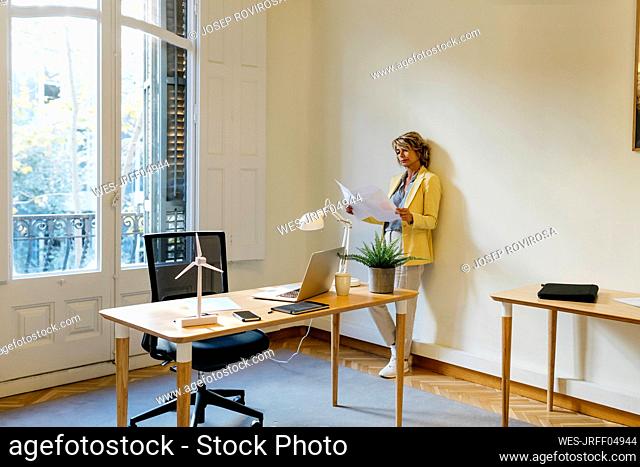 Businesswoman examining blueprint while standing against wall in office