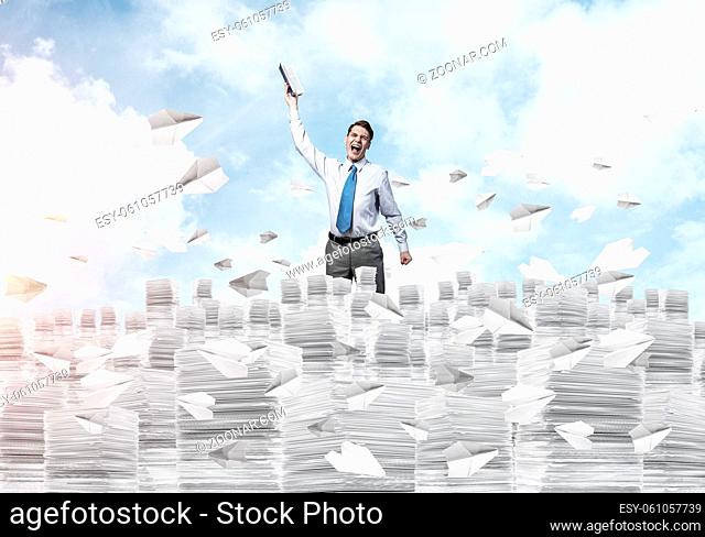 Businessman keeping hand with book up while standing among flying paper planes with cloudly skyscape on background. Mixed media