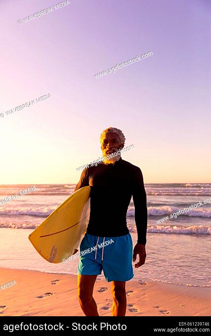 Bearded african american senior man with surfboard standing against sea and clear sky at sunset
