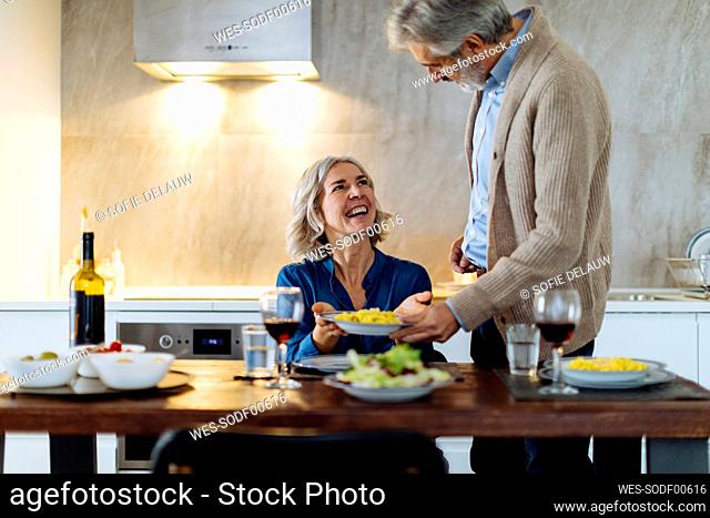 Mature man serving dinner to happy wife in kitchen at home