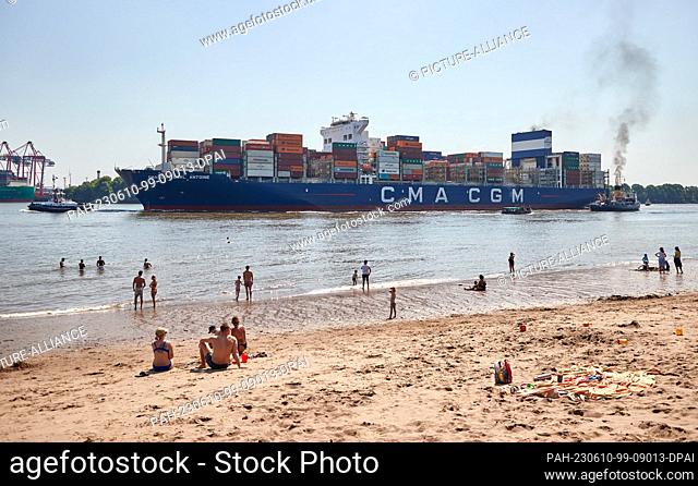 10 June 2023, Hamburg: People enjoying the sun on the Elbe beach at Övelgönne while the container ship ""CMA GM Carl Antoine"" sails upstream in the background