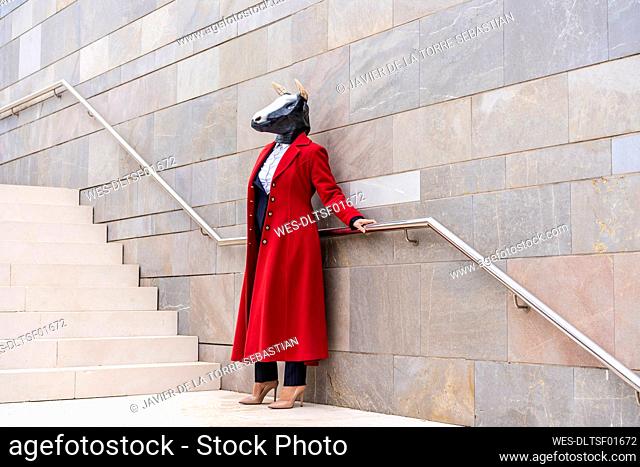 Woman wearing red overcoat and bull mask holding railing while standing near staircase