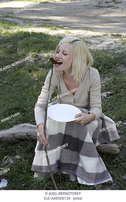 Woman at campsite eating a hotdog off a stick and smiling