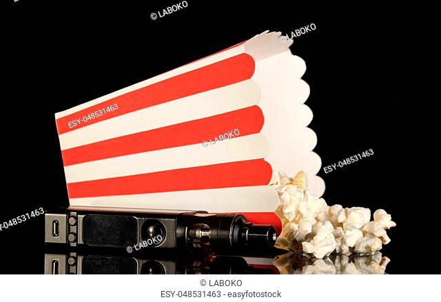 Popcorn fell out of the striped box next to electronic cigarette isolated on black background