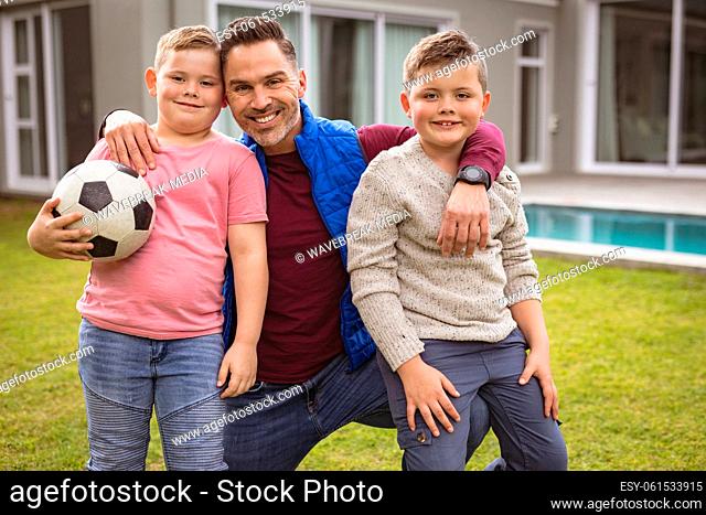 Portrait of caucasian father and two sons smiling while holding football in the garden