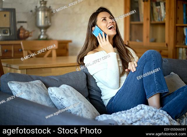 beautiful young smiling woman in white sweater talking on the phone on a gray sofa