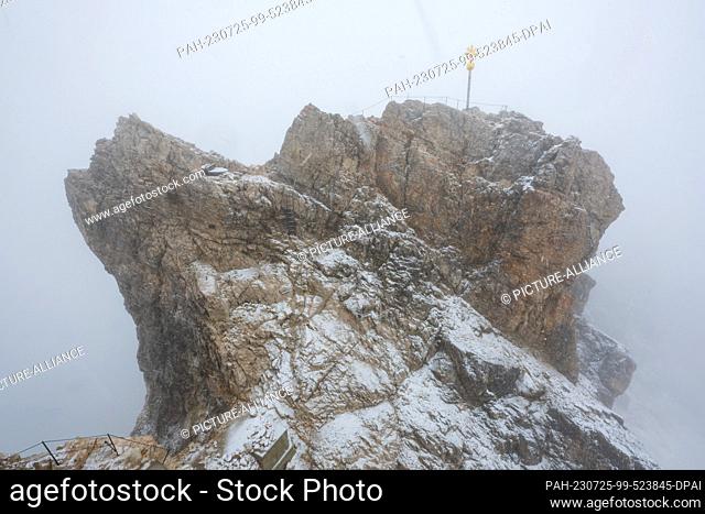 25 July 2023, Bavaria, Garmisch-Partenkirchen: Clouds of fog envelop the snow-covered viewing platform with the summit cross in driving snow on the Zugspitze...