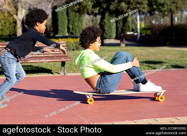 Brother running behind sister sitting on skateboard while playing at park