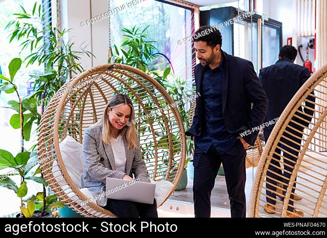 Businesswoman sitting with laptop in hanging chair talking to businessman