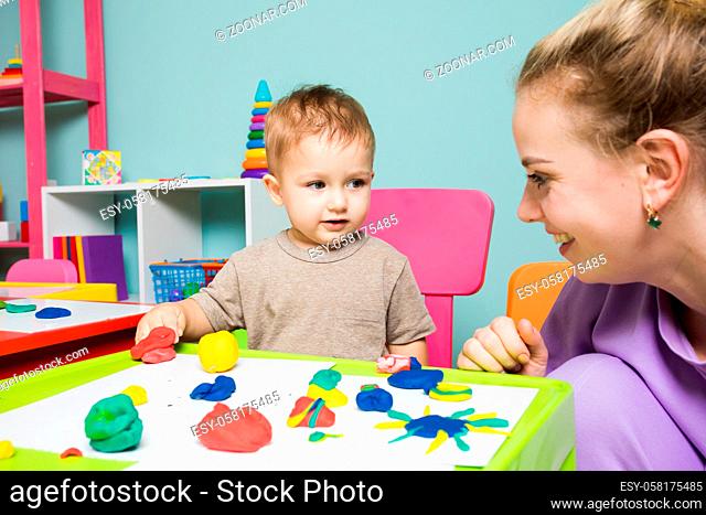 The toddler boy and woman caregiver are looking at each other. The boy sculpts uses plasticine with the help of a tutor