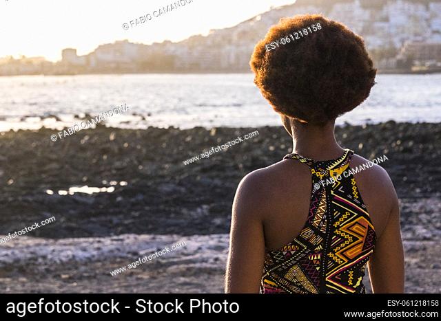 Back view of afro ethnic woman with traditional dress and african hair style looking at the ocean and sunset - black female people enjoy travel and outdoor...