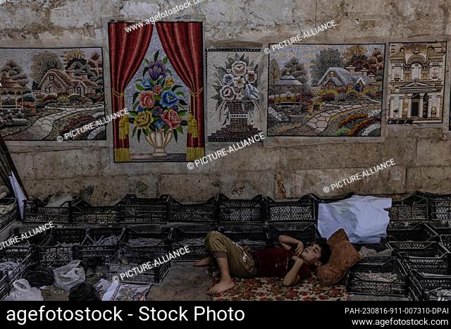 15 August 2023, Syria, Idlib: A Syrian worker sleeps as mosaic paintings hanged in a workshop in Idlib Governorate. Idlib is famous for its mosaic art