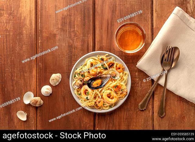 Seafood pasta. Tagliolini with mussels, shrimps, clams and squid rings, with a glass of white wine, shot from the top on a dark rustic wooden background with a...