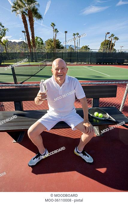 Portrait of happy senior male tennis player with water bottle relaxing on court