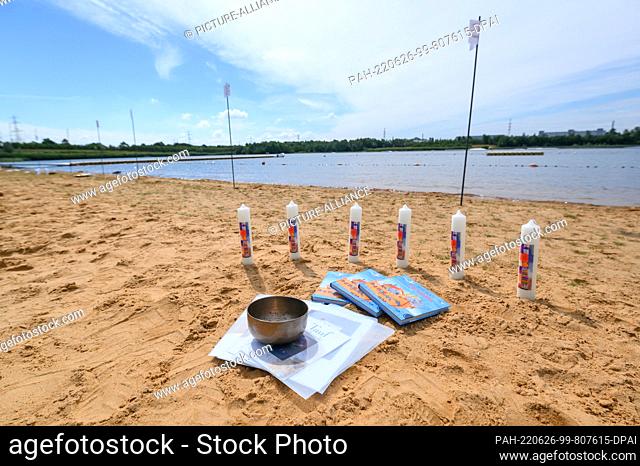 26 June 2022, Schleswig-Holstein, Norderstedt: Baptismal candles, books and a metal bowl stand on the beach on the shore of the Stadtparksee before the baptism...