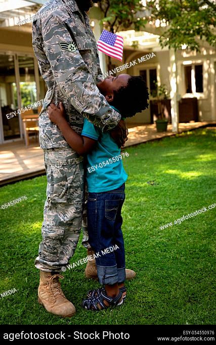 SIde view close up of a young adult African American male soldier in the garden outside his home, embracing his young son