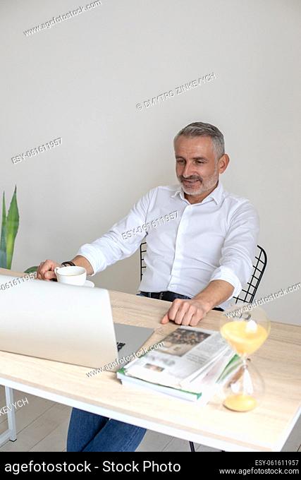 Interesting here. Middle-aged man with coffee looking interestedly into laptop sitting leaning back in chair at table in room