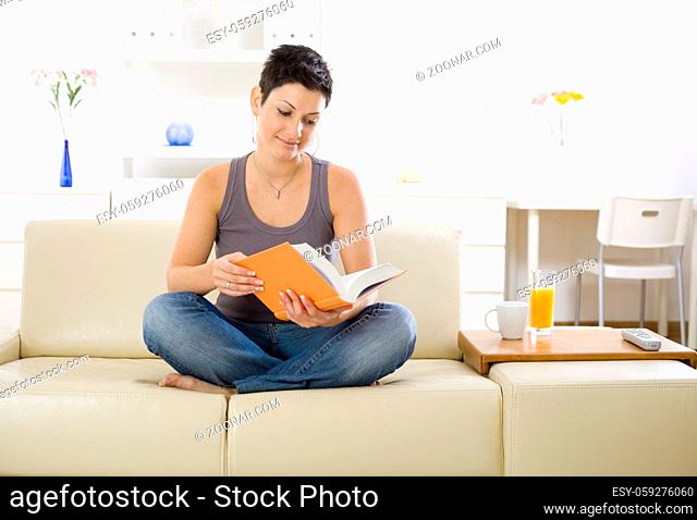 Female student sitting on sofa at home, reading book