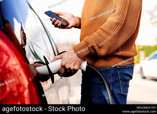 Man with smart phone plugging in electric plug at vehicle charging station