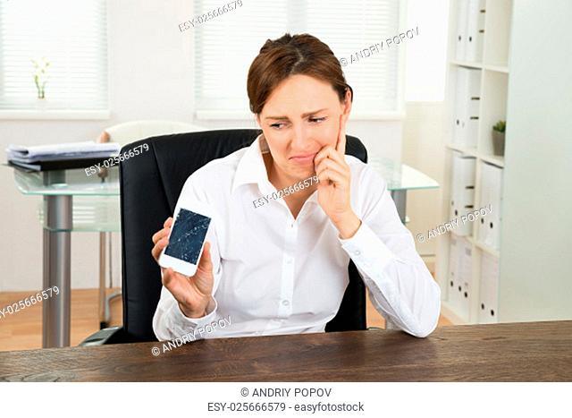 Unhappy Young Businesswoman Looking At Broken Mobile Phone In Office
