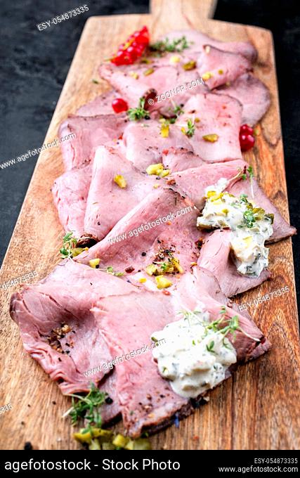 Traditional lunch meat with sliced cold cuts roast beef decorated with remoulade sauce and red ribes as top view on a cutting board