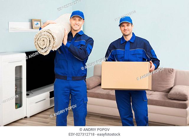 Portrait Of A Two Male Movers Carrying Rolled Carpet And Cardboard Box In House