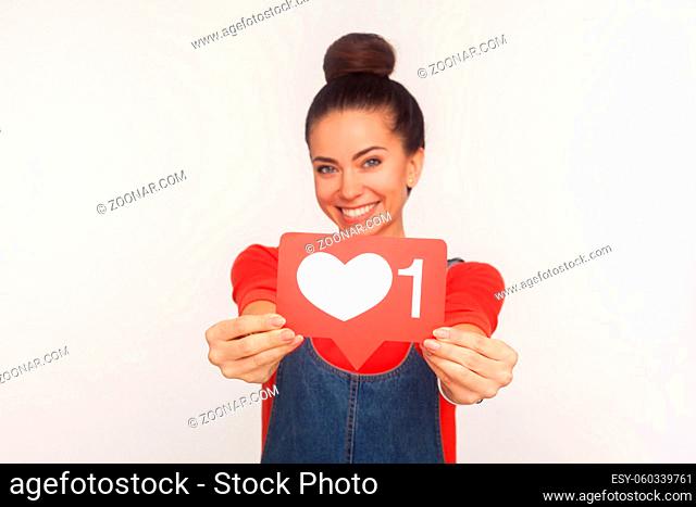 Like and follow interesting content. Portrait of charming positive trendy girl with hair bun in denim overalls showing social media heart icon to camera and...