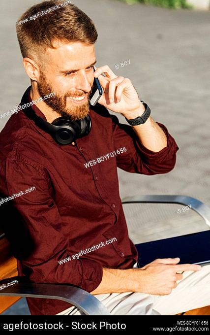Entrepreneur smiling while talking on mobile phone sitting on bench during sunny day
