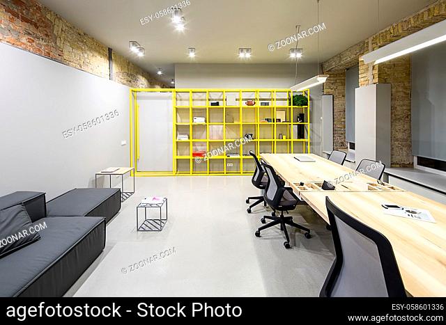 Office in loft style with gray and brick walls. There are glowing lamps, dark sofa with pillow, wooden tables with chairs, small metal tables