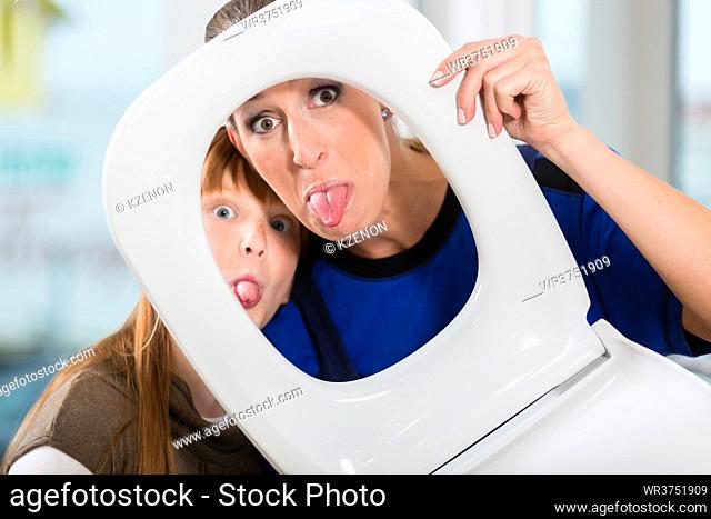 Funny portrait of a cheerful woman and her cute daughter smiling and looking at camera through a white toilet seat in a sanitary ware shop