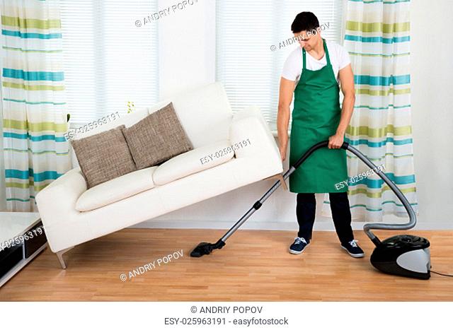 Full length of man lifting couch while cleaning hardwood floor with vacuum cleaner at home