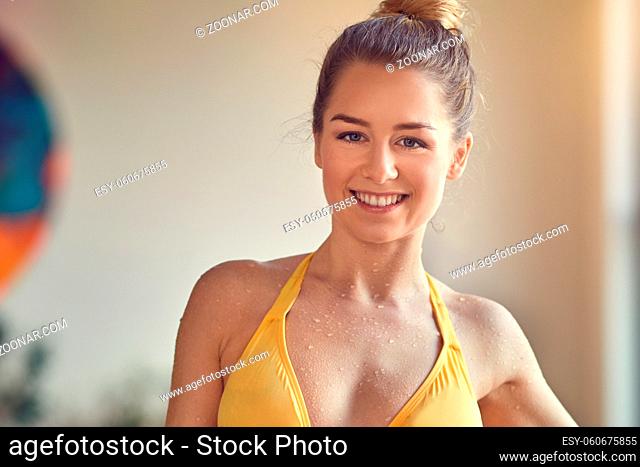 Beautiful young sporty woman standing in swimming pool in yellow two-piece swimsuit, looking away and smiling
