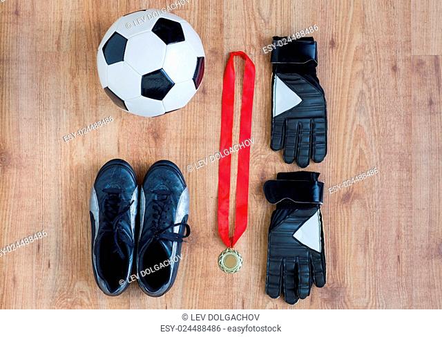 sport, achievement, championship and success concept - close up of soccer ball, football boots and goalkeeper gloves with golden medal on wooden background