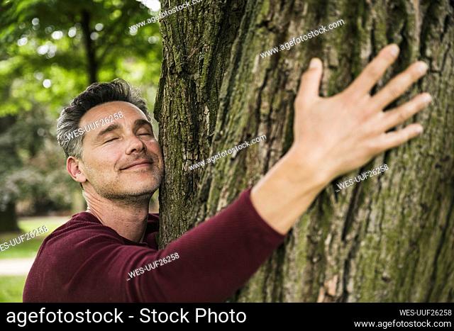 Smiling man with eyes closed hugging tree at park