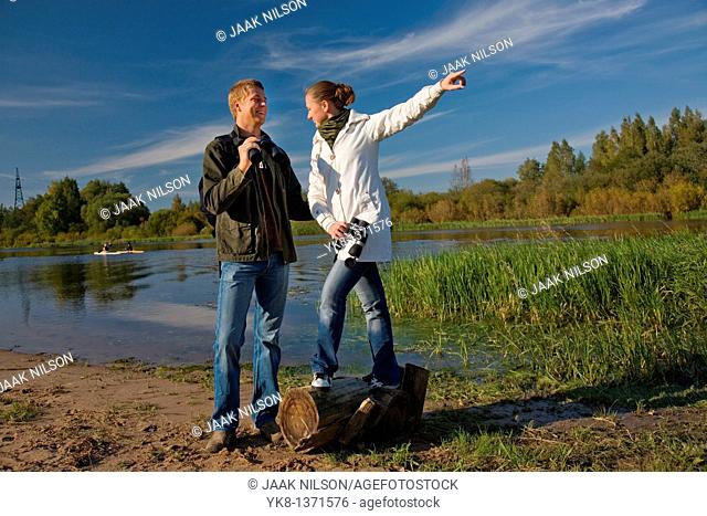 Young couple birdwatching with binoculars by river water pointing away