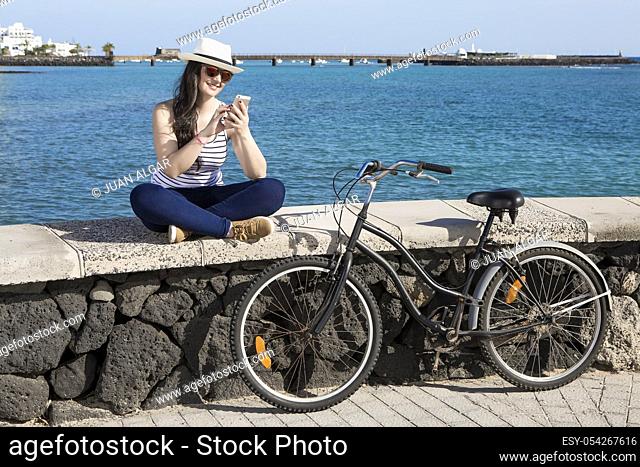 Young smiley woman in hat and sunglasses using device while sitting on quay in Lanzarote, Gran Canaria