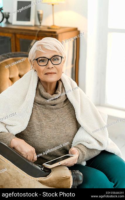 Elegant Old lady Wearing Glasses Is Sitting On The Leather Arm-chair And Holding A Tablet In Her Hand. The Woman Is Wrapped In A Woolen Plaid
