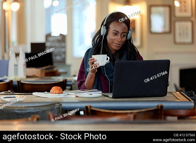 Young woman with headphones drinking coffee at laptop in cafe