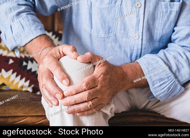 Close up of senior old man facing knee problem, sitting on sofa holding knee at home. Elderly man suffering from severe knee ache sitting at home