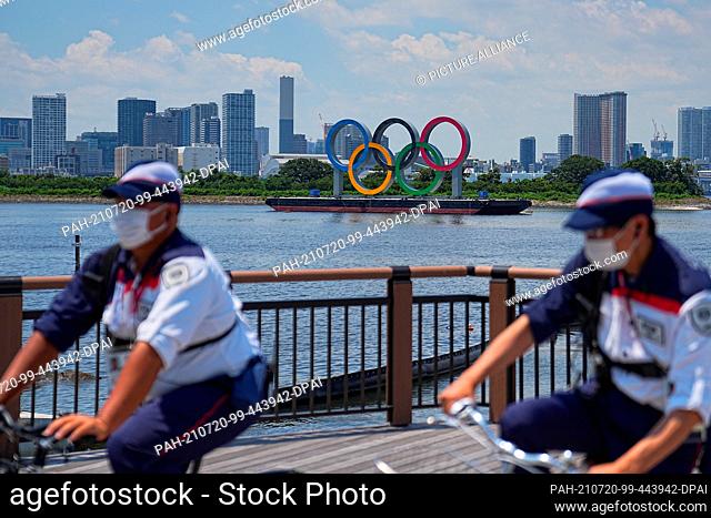 20 July 2021, Japan, Tokio: Two security guards ride bicycles in front of the Olympic rings. The rings are on a pontoon in Tokyo Bay
