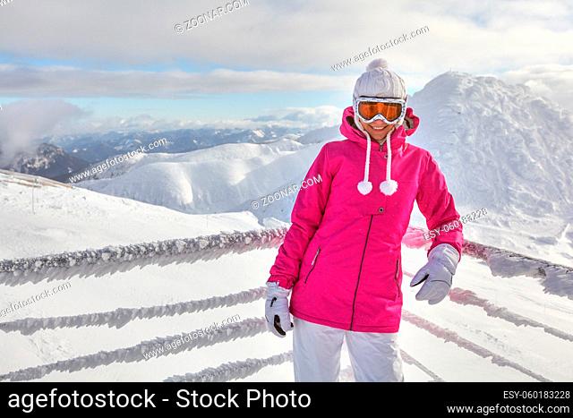 Young woman in pink jacket, wearing ski goggles, leaning on snow covered fence, smiling, white mountains in background