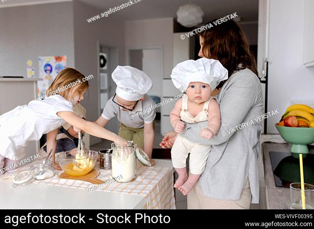 Mother carrying boy with children preparing dough in kitchen